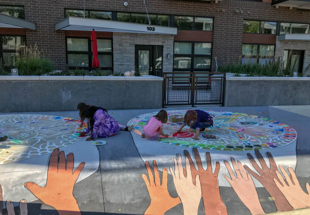 Three people, including two children, kneel on a colorful chalk mandala drawn on a public sidewalk. In the foreground are painted raised hands, and an apartment building is visible in the background.