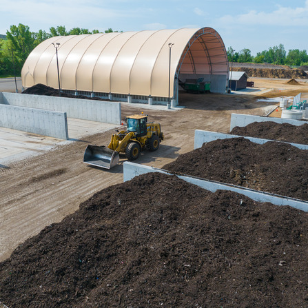 Front-end loader moving compost material in an outdoor composting facility with concrete bays and a covered storage structure.