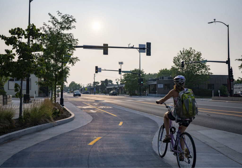 A person wearing a helmet and backpack rides a bicycle on a dedicated bike path beside a city street with traffic lights, trees, and buildings under a hazy sky.