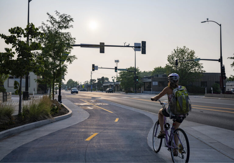 A person wearing a helmet and backpack rides a bicycle on a dedicated bike path beside a city street with traffic lights, trees, and buildings under a hazy sky.