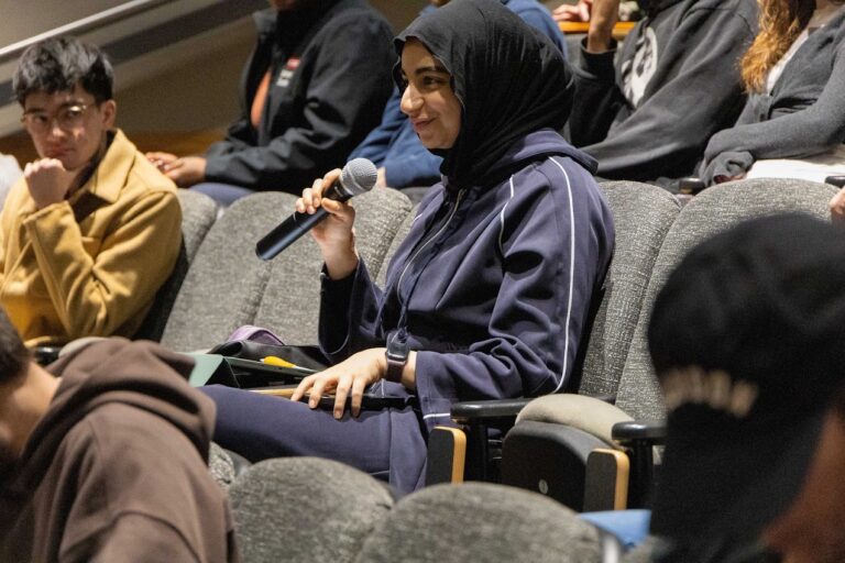 A woman wearing a dark hijab and tracksuit is seated in an auditorium, smiling and holding a microphone, appearing ready to speak about career paths for students. Other people are seated around her, listening attentively.