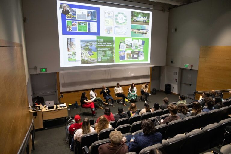 A group of five panelists speak on stage in a lecture hall to an audience. A large screen behind them displays slides featuring career paths for students and information about environmental or community initiatives.