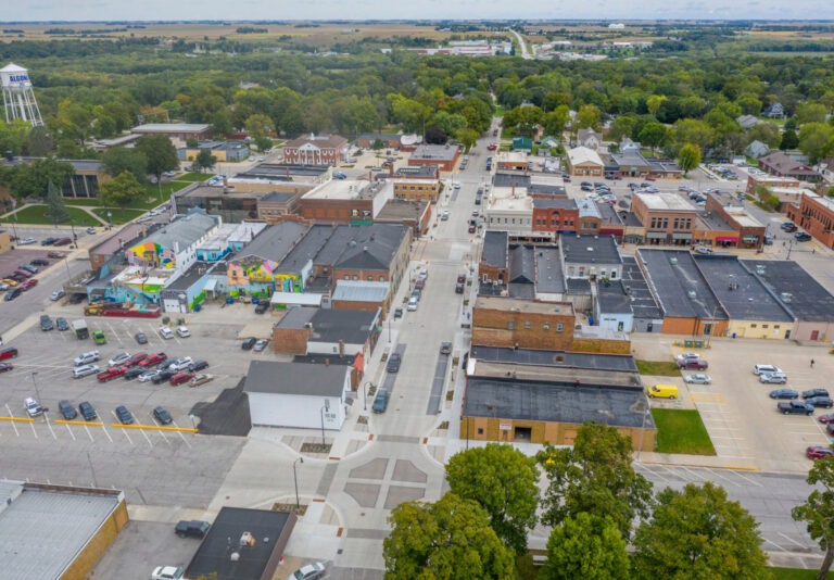 Aerial view of a small town with tree-lined streets, brick buildings, a water tower, parked cars, and vibrant murals on some walls; farmland and greenery surround the town.