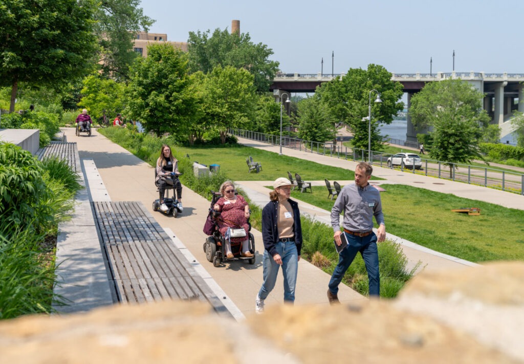 People using power wheelchairs and others walking along a wide, accessible path in a green park on a sunny day. Trees, benches, and a bridge are visible in the background.