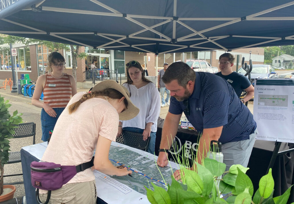 A group of people stands around a table under a canopy, examining a large map and discussing. One person points at the map while others look on attentively. There are plants and informational posters on the table.