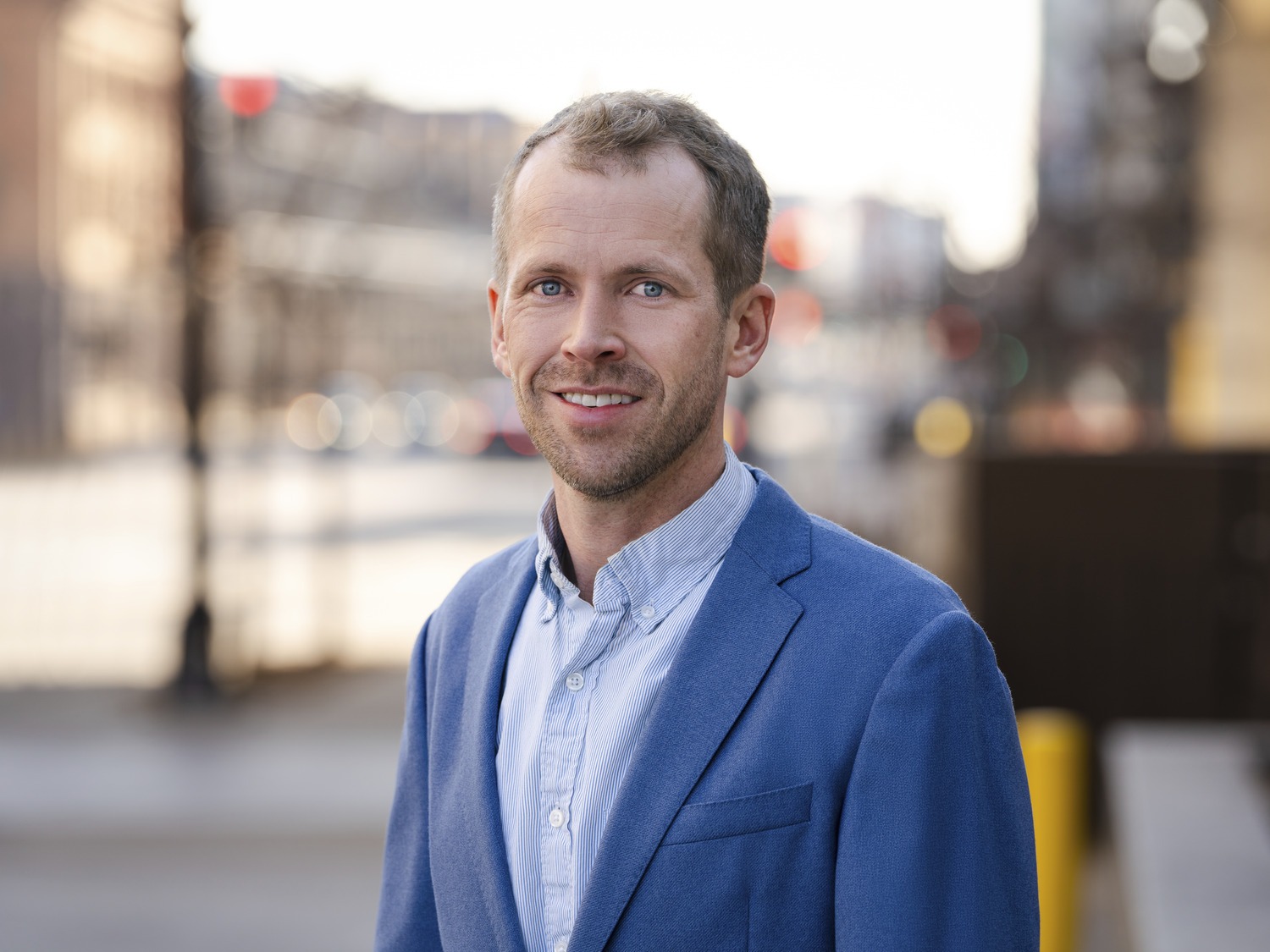 A man with short light brown hair and a trimmed beard, wearing a blue blazer over a light blue shirt, stands outdoors on a city street.