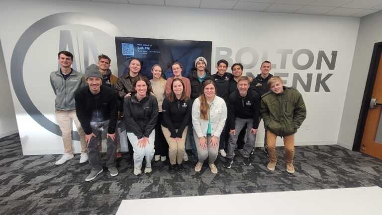 A group of sixteen people pose and smile for a photo in an office lobby with Bolton & Menk on the wall behind them, showcasing career paths for students. Most are standing, with four kneeling in front. The group appears cheerful and relaxed.