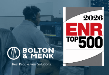 A man wearing headphones works at a computer in an office cubicle. Overlaid logos include Bolton & Menk with the text Real People. Real Solutions. and 2026 ENR Top 500.