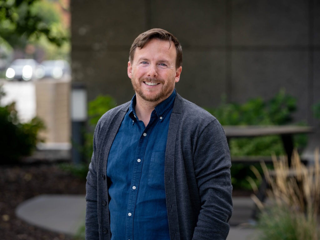 A man with light brown hair and a beard, wearing a blue shirt and gray cardigan, stands outdoors smiling.
