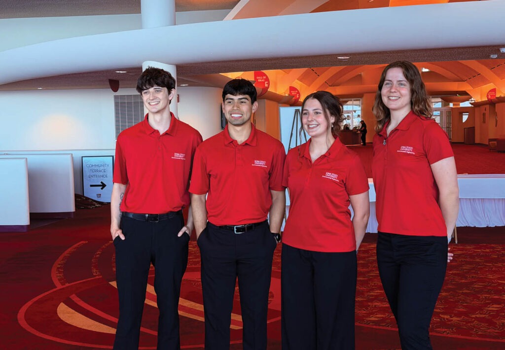 Four young adults wearing matching red collared shirts and black pants stand side by side, smiling, in a brightly lit indoor setting with red carpet and white walls.