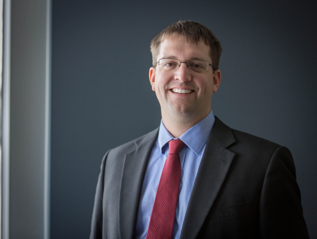 A man wearing glasses, a dark suit, light blue shirt, and red tie stands indoors, smiling in front of a plain dark background.