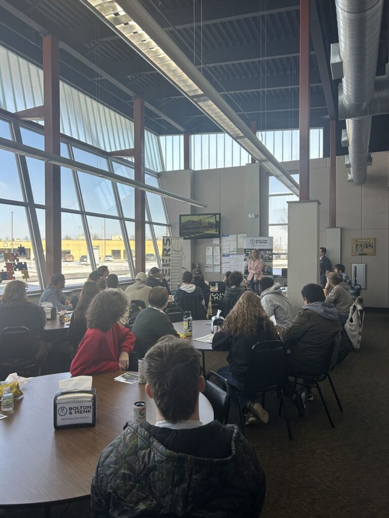 A group of people sit at round tables in a modern, sunlit room with large windows, listening to a speaker discuss career paths for students near a screen. Lunch items and beverages are on the tables.
