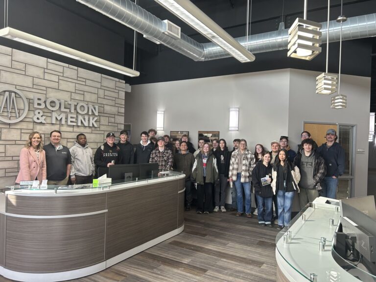 A large group of people, exploring career paths for students, poses for a photo inside the Bolton & Menk office lobby, standing behind and beside a curved reception desk surrounded by modern decor with exposed ducts and a stone accent wall.