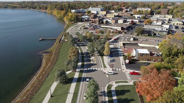 Aerial view of a small town with buildings, roads, parked cars, and a waterfront with a dock. Trees, some with autumn foliage, line the curved shoreline and streets. A park with walking paths borders the water.