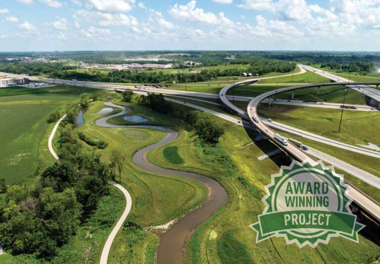 Aerial view of a highway interchange with curving roads, green fields, and a winding stream. A badge in the foreground reads Award Winning Project. Blue sky with scattered clouds above.
