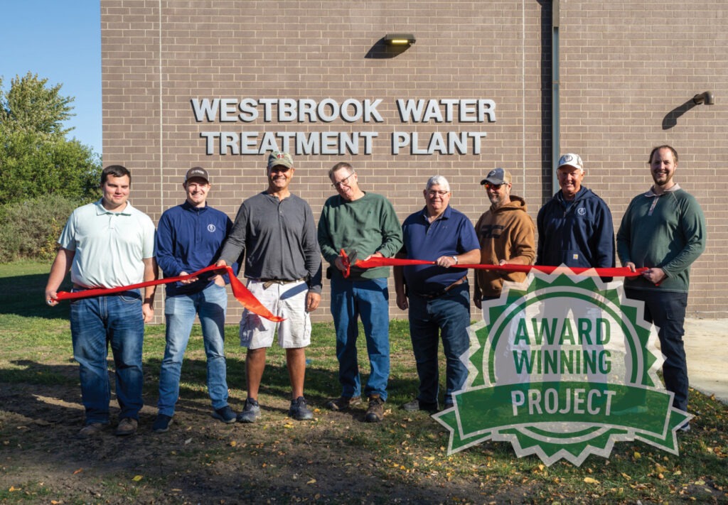Eight people stand in front of the Westbrook Water Treatment Plant holding a red ribbon. A badge in the lower right corner reads Award Winning Project with AQUARIUS Recognition. The group is outside, surrounded by sunlight and greenery.