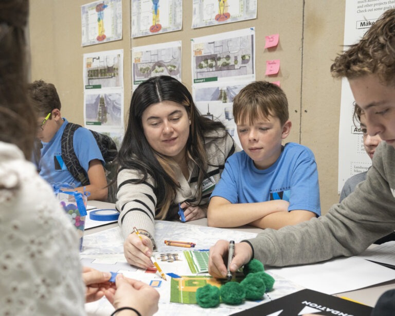 A woman and several children sit around a table working on a hands-on project with papers, models, and markers—exploring ideas related to career paths for students in a focused classroom or workshop setting with diagrams on the wall behind them.
