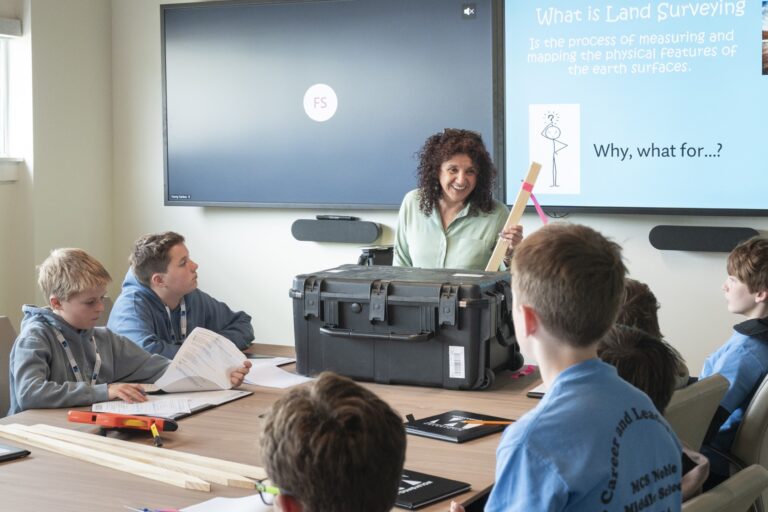 A woman stands at the front of a classroom holding surveying equipment, engaging a group of attentive students seated around a table. A presentation about land surveying and career paths for students is displayed on a screen behind her.