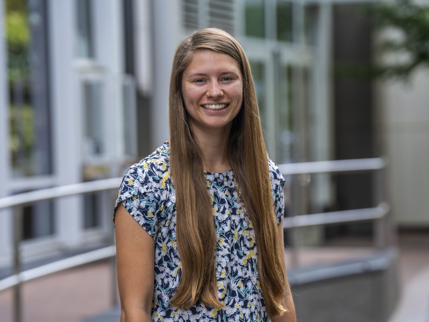 A young woman with long, straight brown hair stands outside a modern building, smiling in a blue patterned short-sleeve blouse. The background features glass windows, a metal railing.