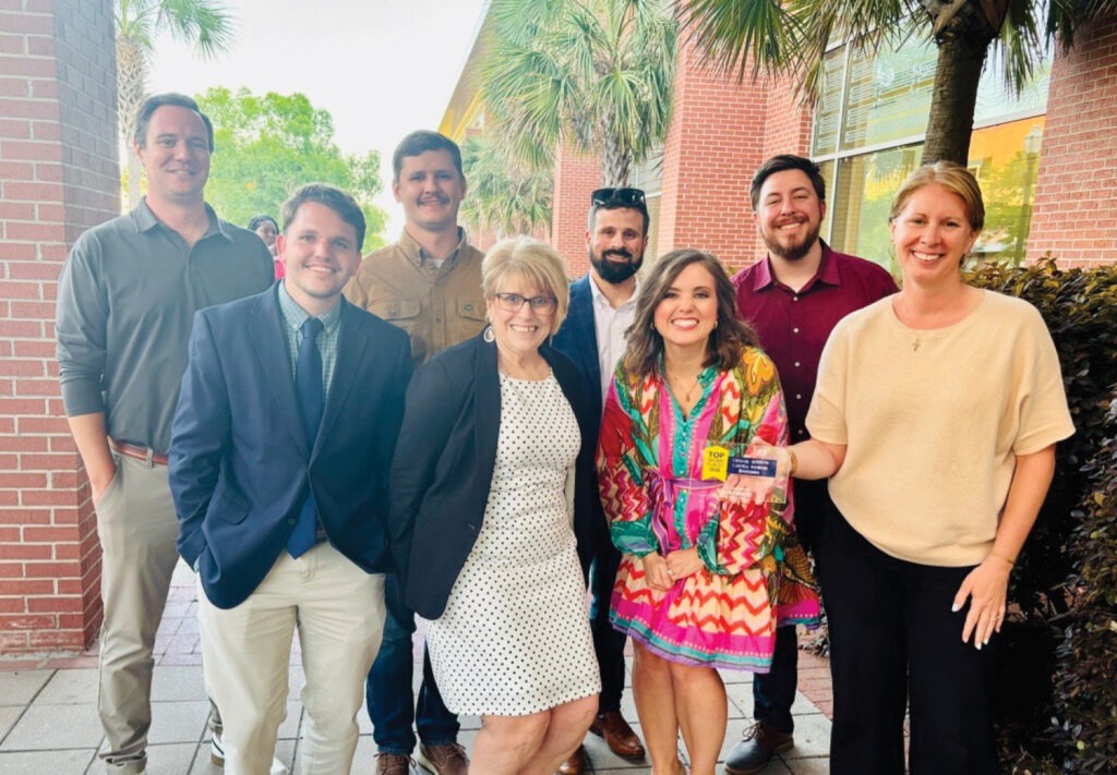 A group of eight adults, dressed in business and casual attire, stand together smiling outdoors near a brick building and palm trees. One woman in a colorful dress holds a card and stands at the front.