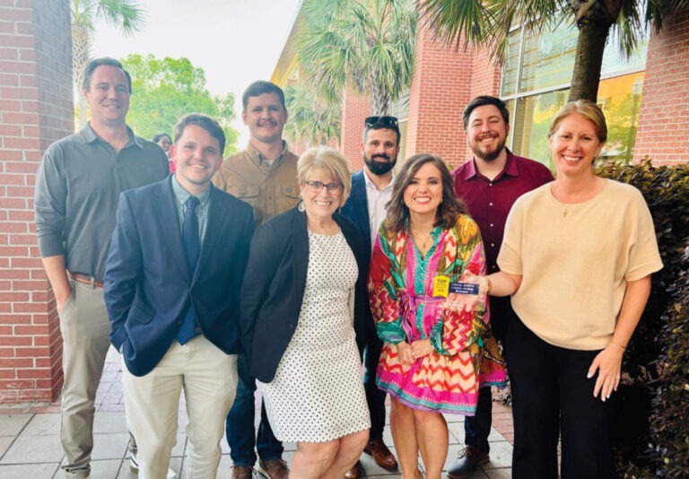 A group of eight adults, dressed in business and casual attire, stand together smiling outdoors near a brick building and palm trees. One woman in a colorful dress holds a card and stands at the front.