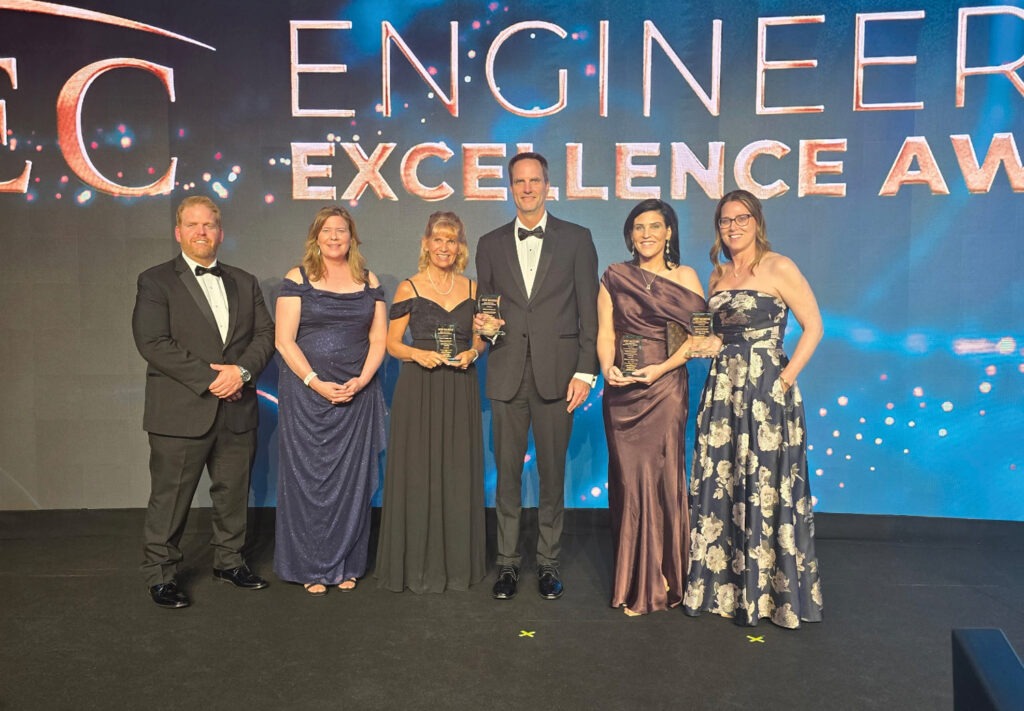 Six people dressed formally stand onstage holding awards in front of a lit backdrop that reads “ENGINEER EXCELLENCE AWARDS,” part of the National Recognition Awards. Three women and one man in the center hold plaques, flanked by two others.