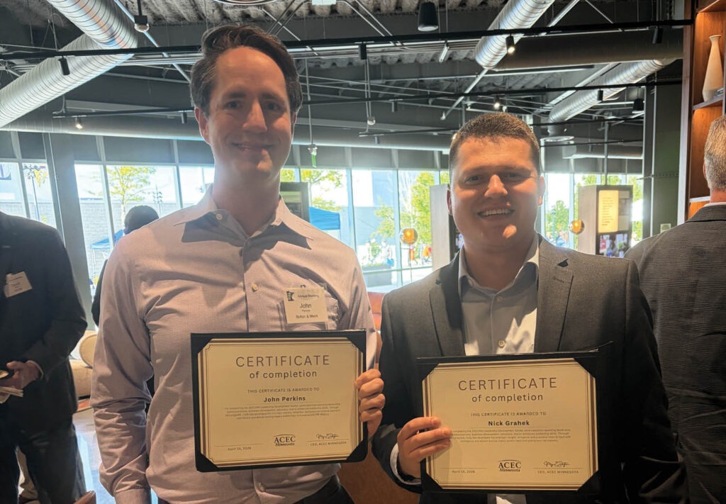 Two men in dress shirts and blazers smile while holding certificates of completion at an indoor event, with other people and large windows in the background.