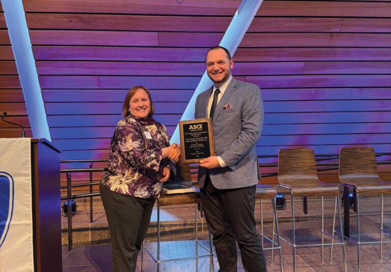 A woman and a man smile as they shake hands on a stage. The man holds a plaque that reads ASCE. Behind them are modern wooden walls, chairs, and a banner partially visible on the left.