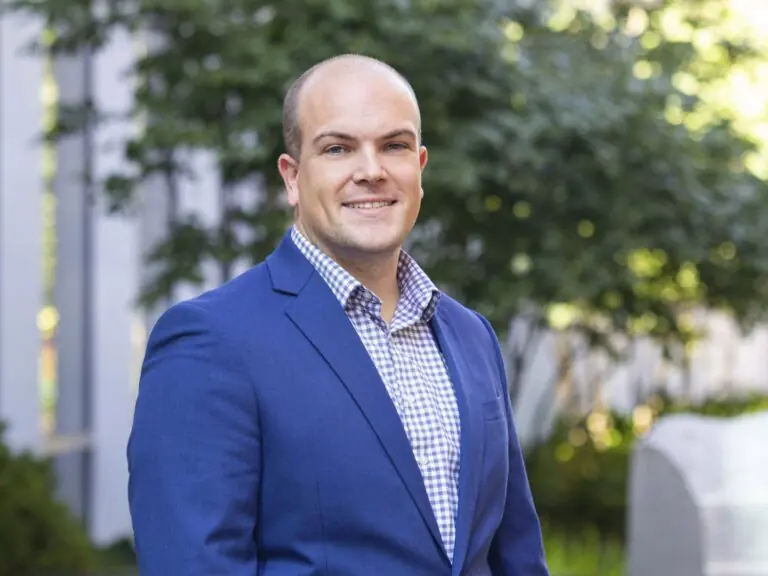 A man in a blue suit jacket and checked shirt smiles while standing outdoors in front of greenery and a building.
