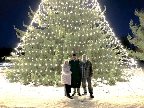 Three people stand in front of a large Christmas tree covered in glowing white lights at night