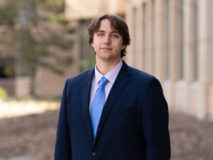 A young man with brown hair in a navy suit, light blue tie, and striped shirt stands outdoors in front of a blurred building and trees, looking at the camera with a neutral expression.