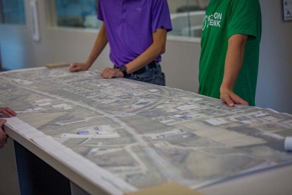 Two people stand at a table, looking at a large, detailed map or blueprint of an urban area spread out before them; only their torsos and arms are visible.