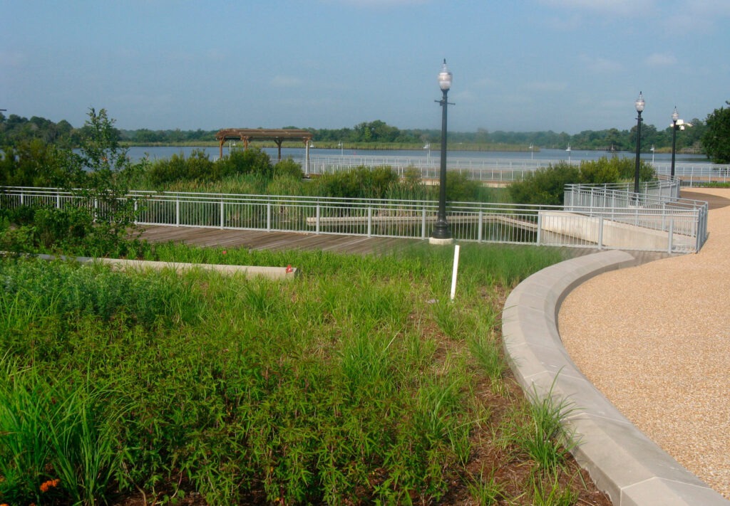 A park with green grass, a curved gravel path, metal railings, wooden deck, lampposts, and a lake in the background under a blue sky highlights effective Green Infrastructure Solutions.