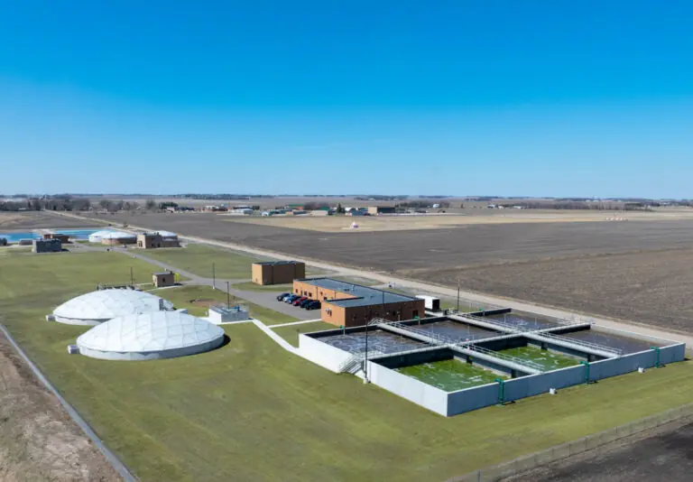 Aerial view of a wastewater treatment plant with large circular covered tanks, rectangular open basins, brick buildings, and surrounding grassy fields next to expansive farmland under a clear blue sky.