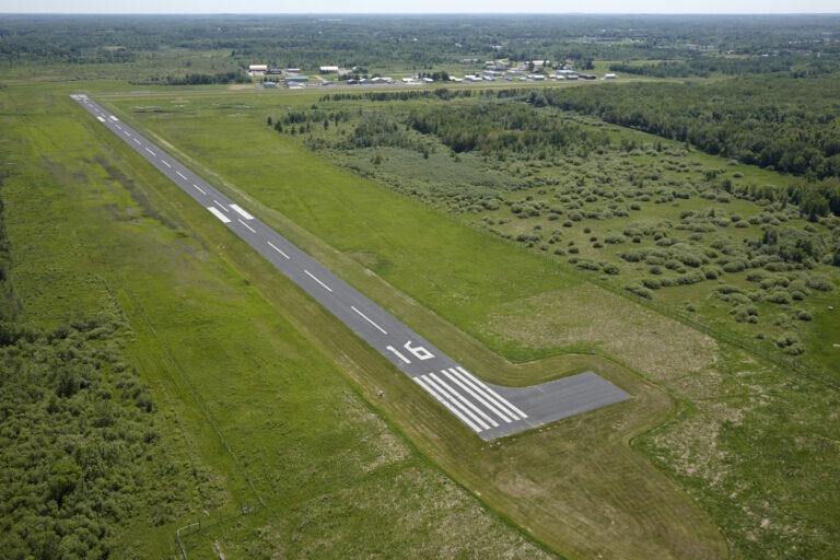 Aerial view of a single runway at a small rural airport, surrounded by green fields, trees, and scattered buildings in the distance under a clear sky.