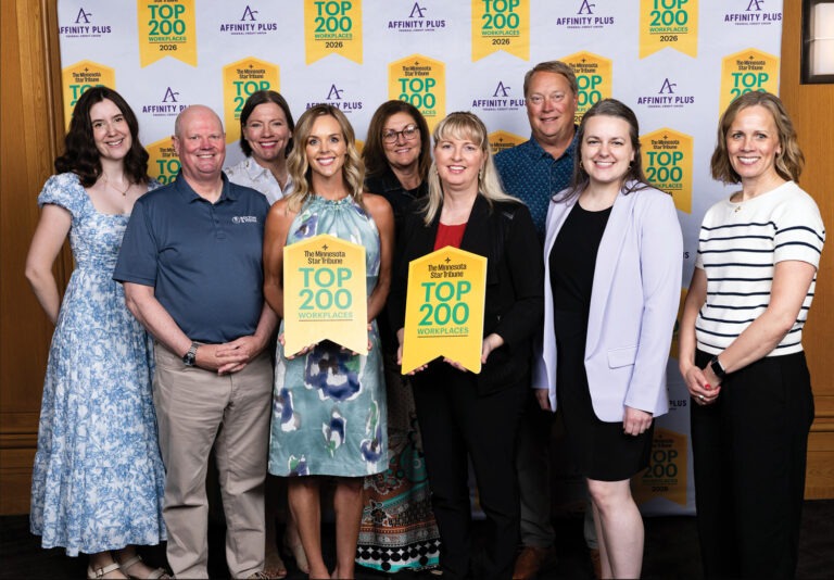 A group of nine people stand smiling in front of a step-and-repeat banner displaying Top 200 Workplaces and Affinity Plus logos. Two people in the front hold yellow Top 200 Workplaces plaques.
