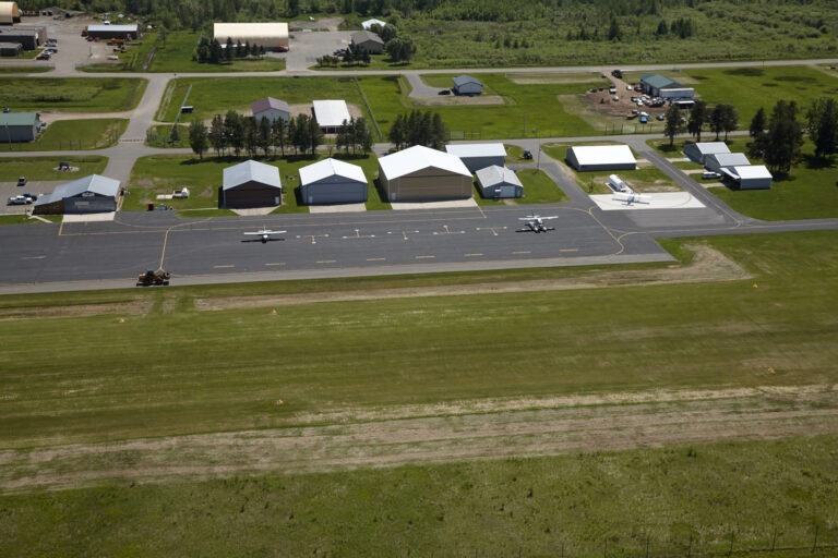 Aerial view of a small airport with several hangars, three small airplanes parked on the tarmac, grassy runways, and surrounding buildings and trees.