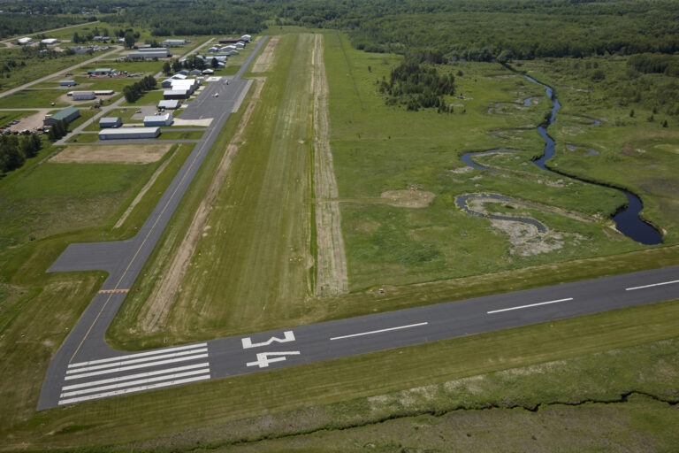 Aerial view of a small airport runway labeled 14, with adjacent taxiways, hangars, parked aircraft, and surrounding grassy fields and a winding creek. Dense forest borders the area in the background.