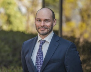 A man in a navy suit and patterned tie smiles outdoors, standing in front of greenery with sunlight filtering through trees in the background.