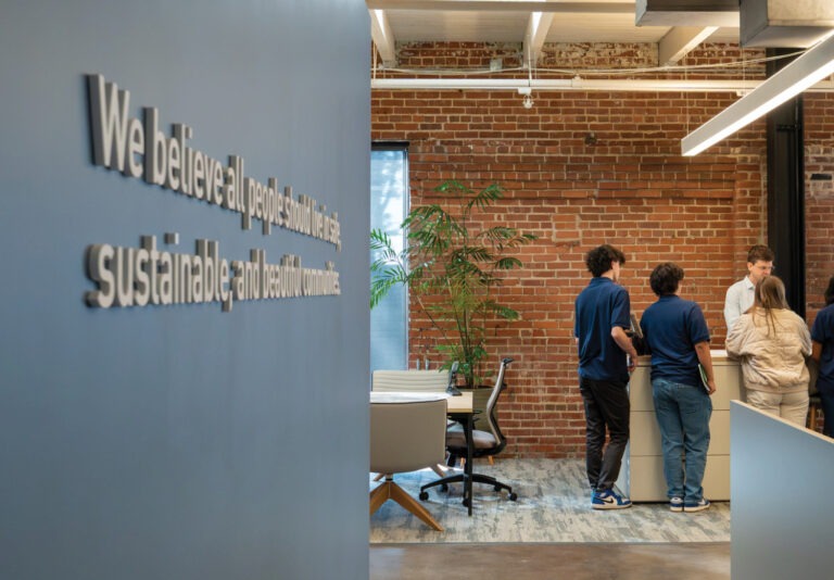 A modern office with exposed brick walls, a sign on the left reading “We believe all people should live in safe, sustainable, and beautiful communities,” and five people conversing near desks and plants.