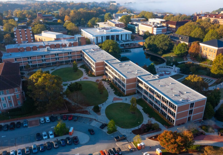 Aerial view of a university campus with modern brick buildings, green lawns, trees with autumn foliage, parked cars, and a reflective pond surrounded by walkways on a clear morning.