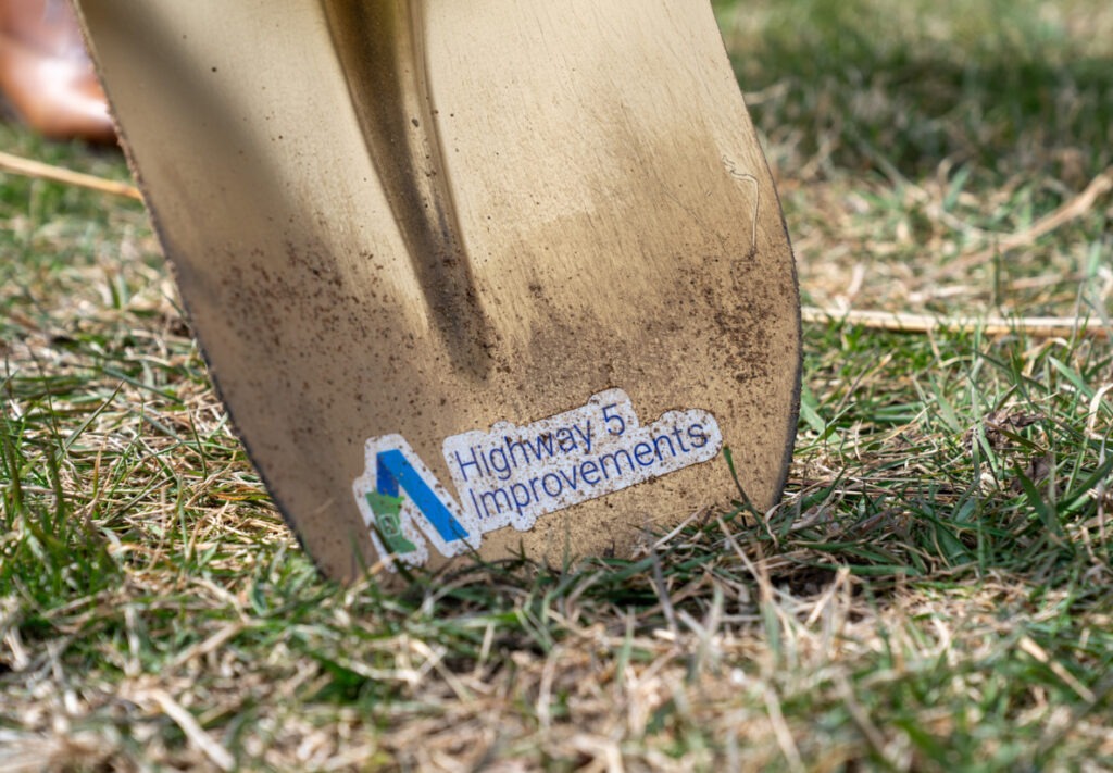 A close-up of a shovel blade with a sticker that reads Highway 5 Improvements pressed into grassy soil.