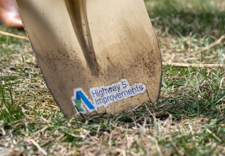 A close-up of a shovel blade with a sticker that reads Highway 5 Improvements pressed into grassy soil.