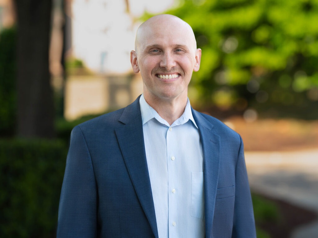 A smiling bald man with a mustache, wears a blue blazer and light blue shirt as he stands outdoors in front of greenery and blurred trees.
