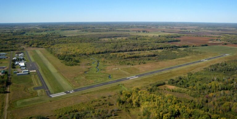 Aerial view of a small rural airport with a single runway supported by airport runway funding, surrounded by green fields and open countryside; several hangars and buildings are visible on the left side.