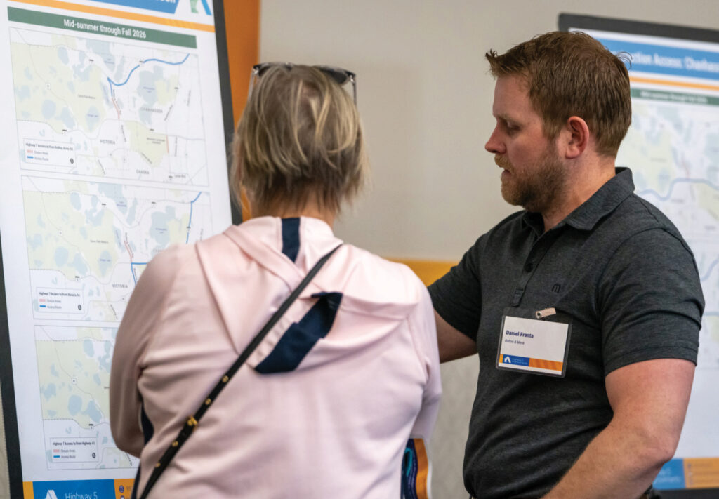 A man and a woman stand in front of a display board with maps and charts, discussing the information. The man points at the board while the woman, with her back to the camera, listens attentively.