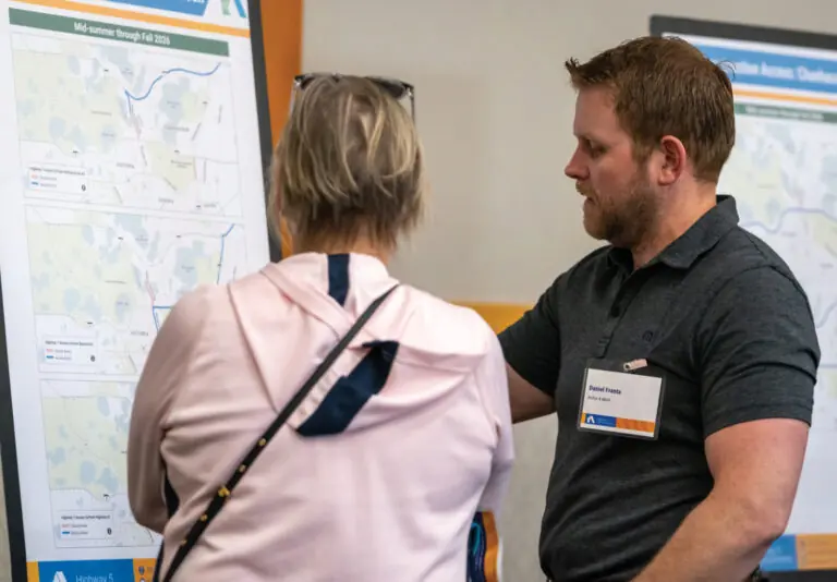 A man and a woman stand in front of a display board with maps and charts, discussing the information. The man points at the board while the woman, with her back to the camera, listens attentively.