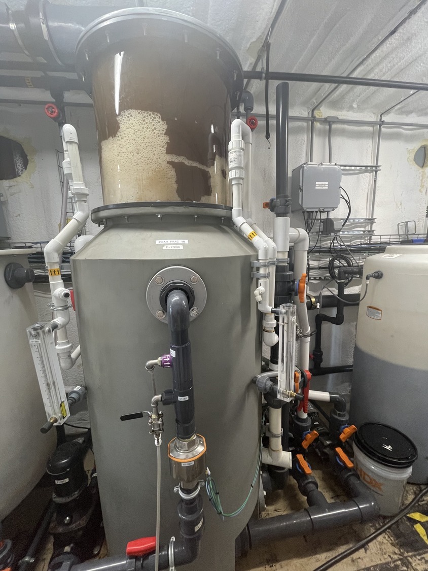 Large cylindrical industrial water filtration unit with pipes and valves, topped with a transparent section containing brown foam. This PFAS Pilot Story system is installed in a utility room with various control panels and containers nearby.