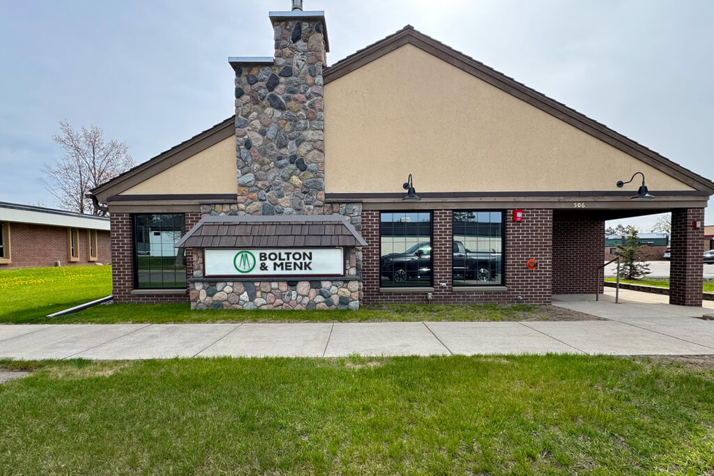 A building with a stone chimney, brick and tan exterior, and a sign reading Bolton & Menk on the lawn, with green grass and sidewalk in front.