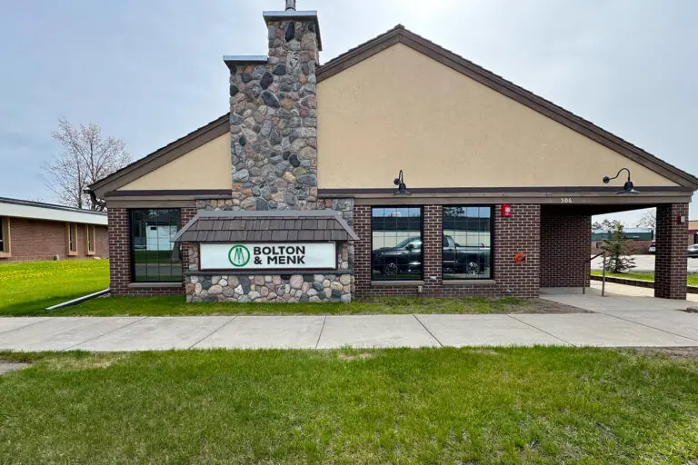 A building with a stone chimney, brick and tan exterior, and a sign reading Bolton & Menk on the lawn, with green grass and sidewalk in front.